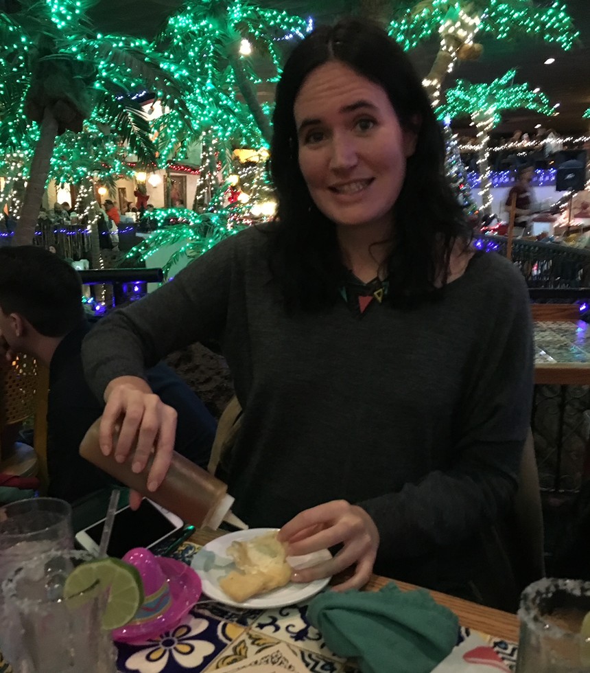 a woman in a sweater holding a squirt bottle of honey in front of a plate of sopaipillas.