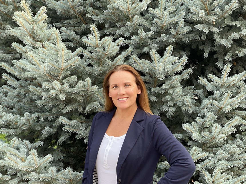 woman with red hair in a blue blazer smiles while posing in front of an evergreen tree.