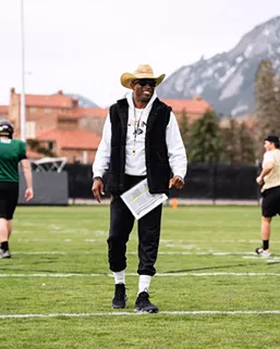 A man in a tan cowboy hat, black vest and white hoodie stands on a football field.