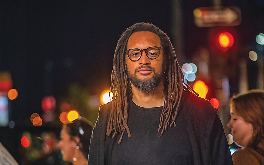 A man with glasses and chest-length dreadlocks looks at the camera with a serious expression.