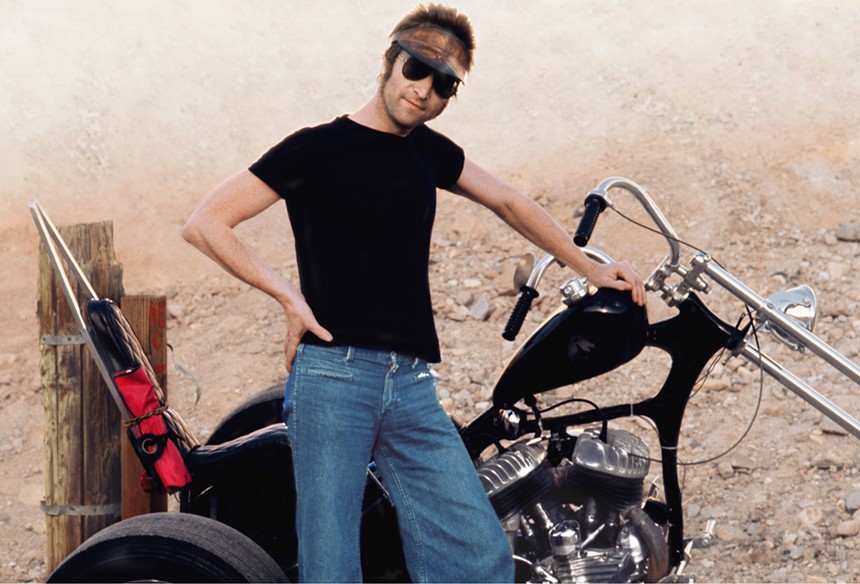 John Lennon in a black t-shirt in jeans posing in front of a motorcycle