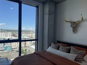 A bed with an orange bedspread sits in front of huge windows.