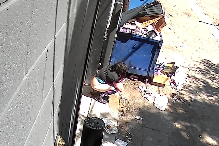 A woman poops behind a dumpster.
