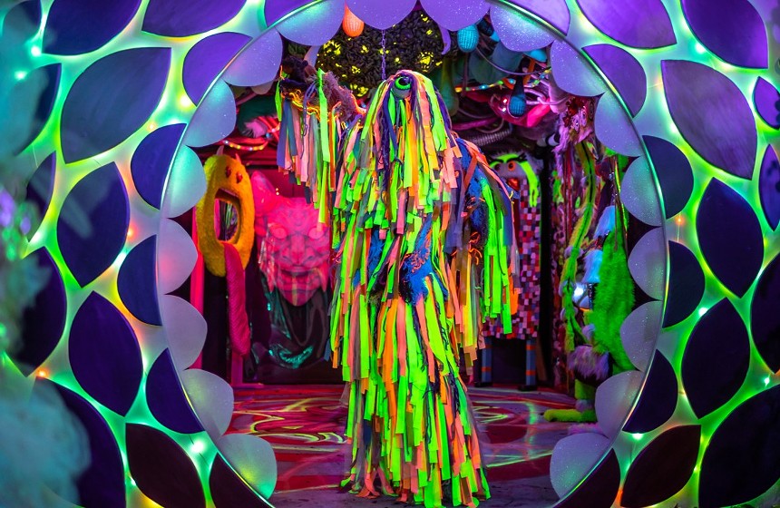 a figure covered in neon fabric stands outside of a psychedelic installation