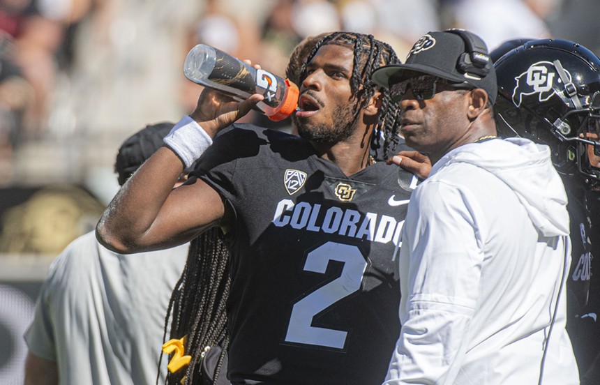 A football player wearing a black jersey drinks from an orange-capped water bottle.