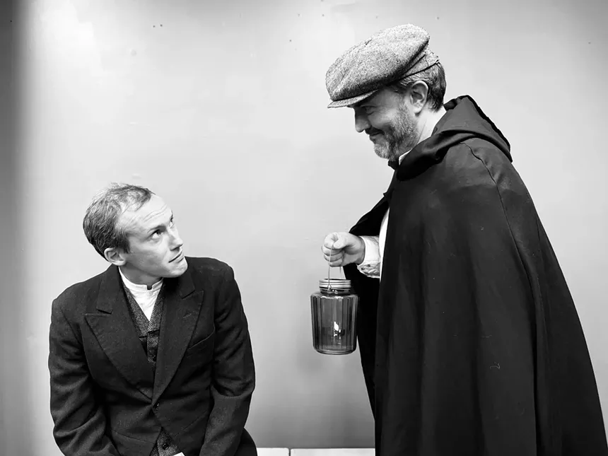 a man in suit sitting looks at a man standing wearing a pageboy hat and black cape while carrying a lantern.