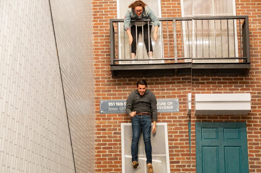 woman standing on porch of brick building while man is hunched over in a grey sweatshirt