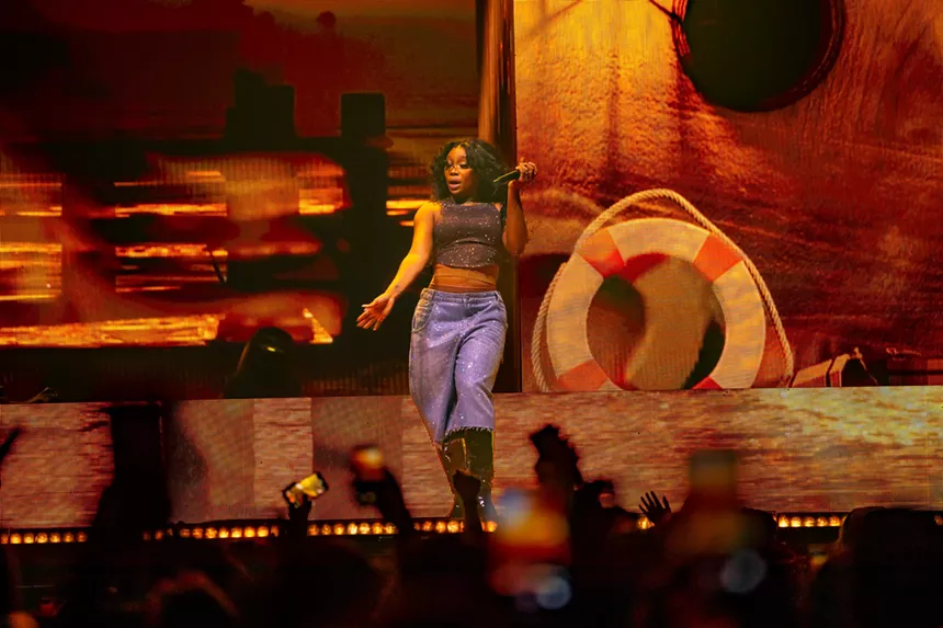woman in blue pants singing into a mic with an orange background