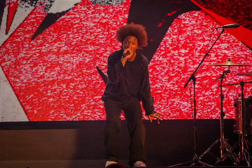 man crouches while singing into a mic with a red background on a screen