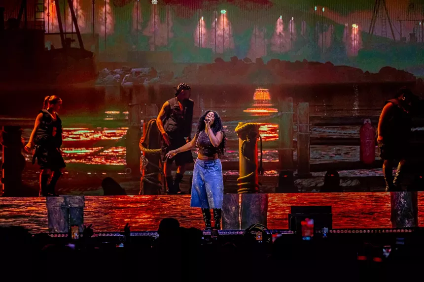 woman in blue pants singing into a mic with an orange background