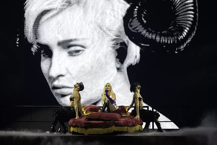 a screen on stage shows a blonde woman with horns, while a blonde woman sings on a bed on stage with two dancers