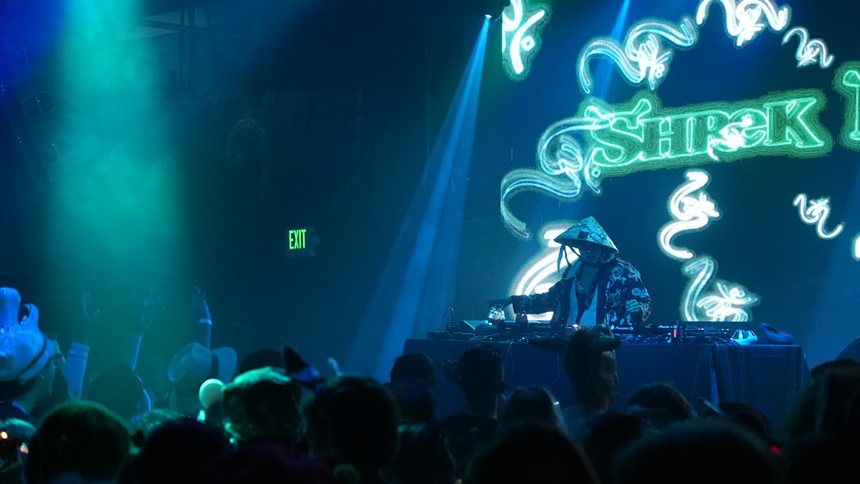 a dj playing to  a crowd while the screen behind him reads "Shrek"