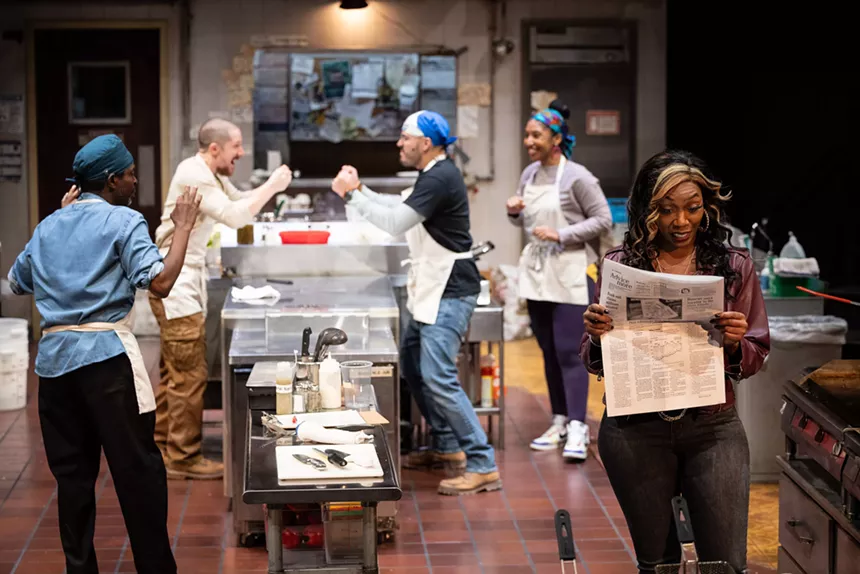 woman to the right reads a newspaper while cooks in the background argue