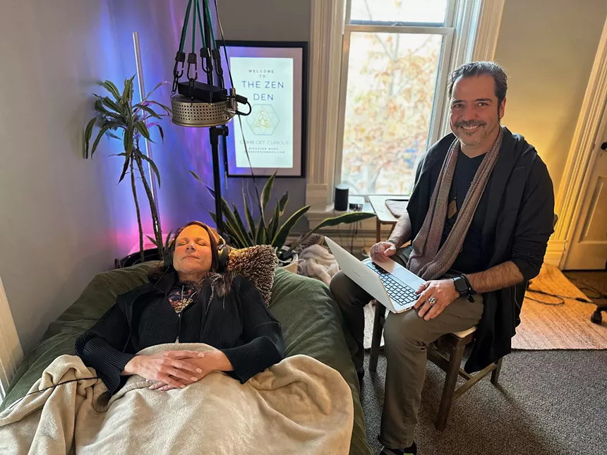 man lies in a bean bag while another sits next to him holding a laptop and smiling