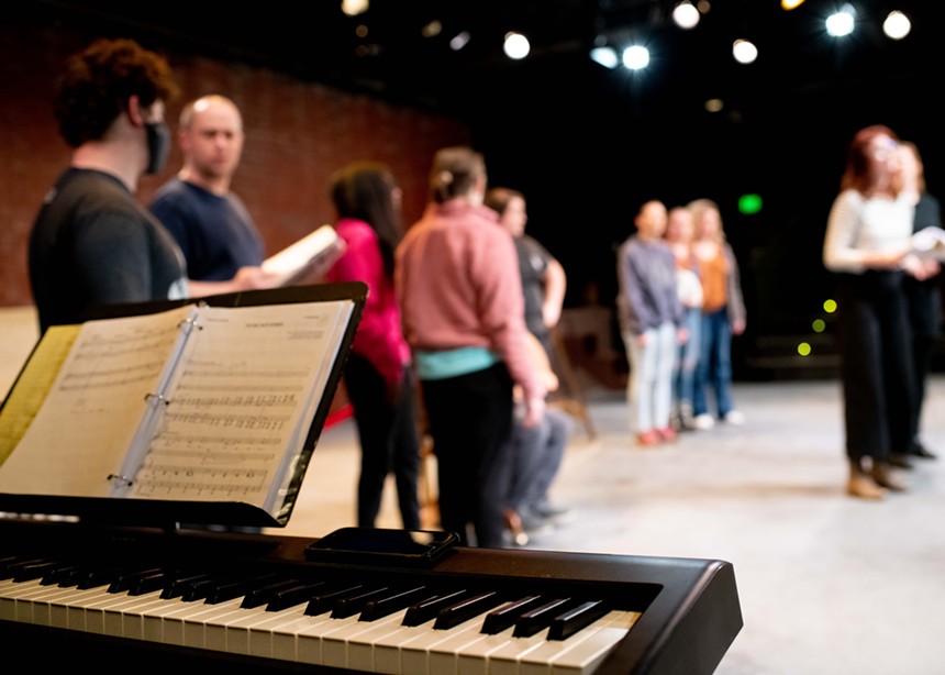 a keyboard piano with actors on stage rehearsing in the background