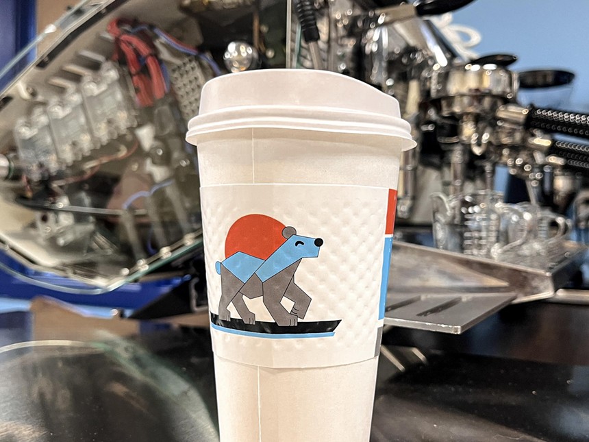 A white coffee cup with a sleeve featuring the Ruzo Coffee logo — a geometric bear backdropped by a red sun