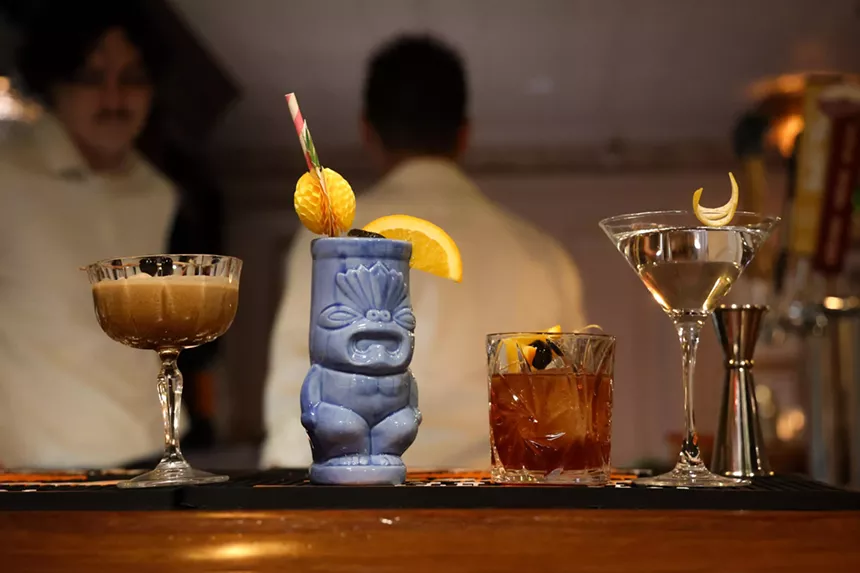 An assortment of four specialty cocktails lined up on a bar in various glassware, with two men in the background