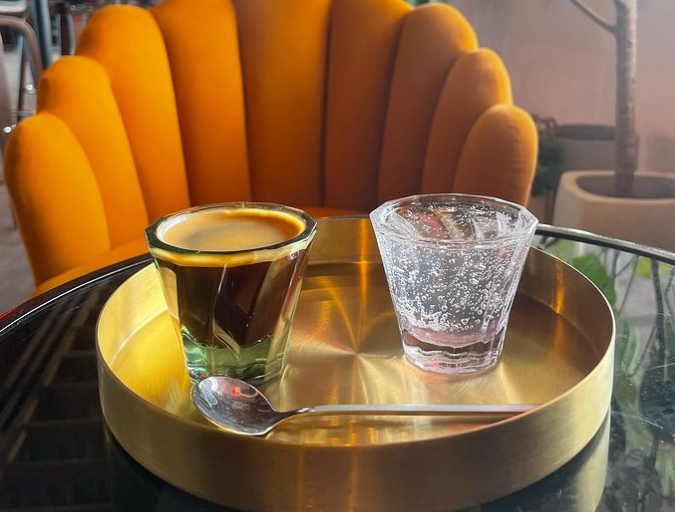 a glass of espresso next to a glass of seltzer in front of an orange chair