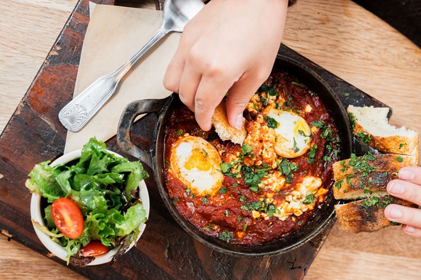 hands dipping bread into tomato sauce with poached eggs