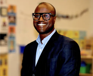 A man smiling in glasses and a blazer with a collared shirt.