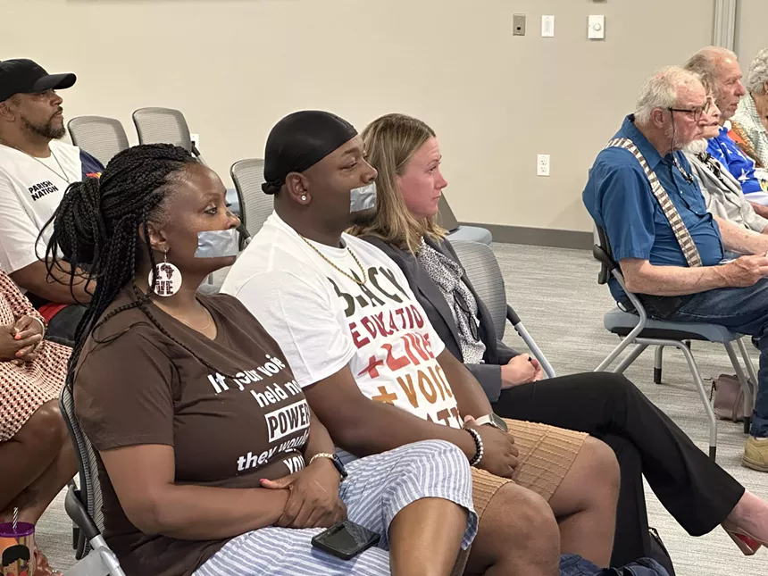 People sitting down during a school board meeting, two of whom have their mouths taped shut in protest of a public comment policy change.