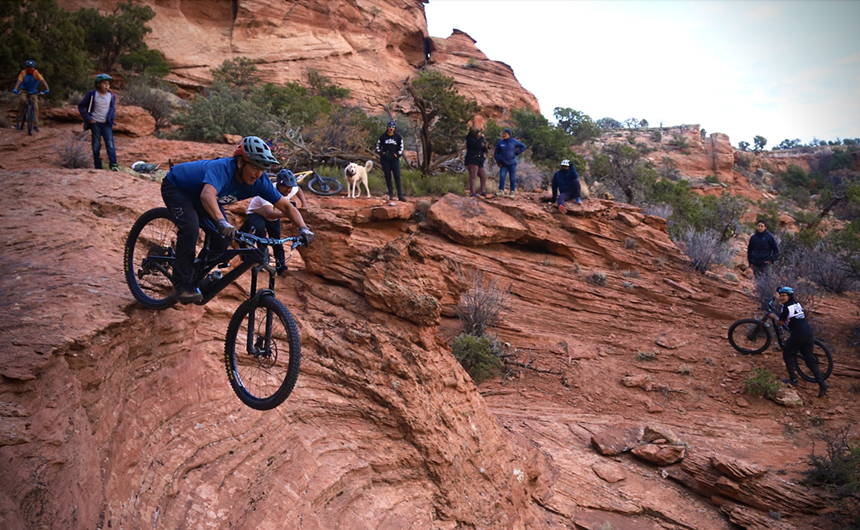 A mountain biker hitting a jump while other riders watch.