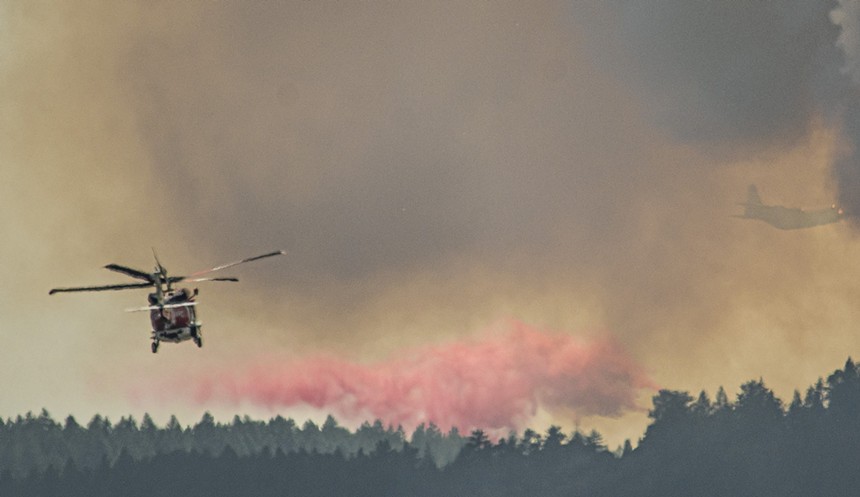 Helicopter flies over trees and red spray from air tanker