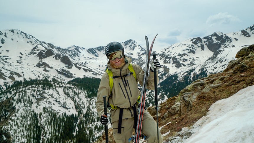 Drew Petersen posed wearing winter gear and holding skis while backdropped by snowy peaks