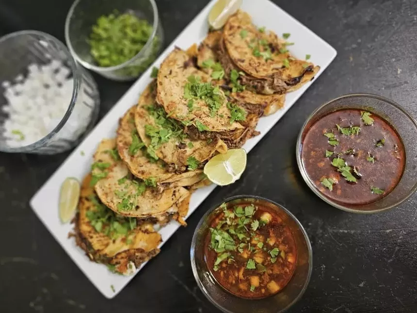 Several birria tacos on a plate next to bowls of broth, cilantro and chopped onions