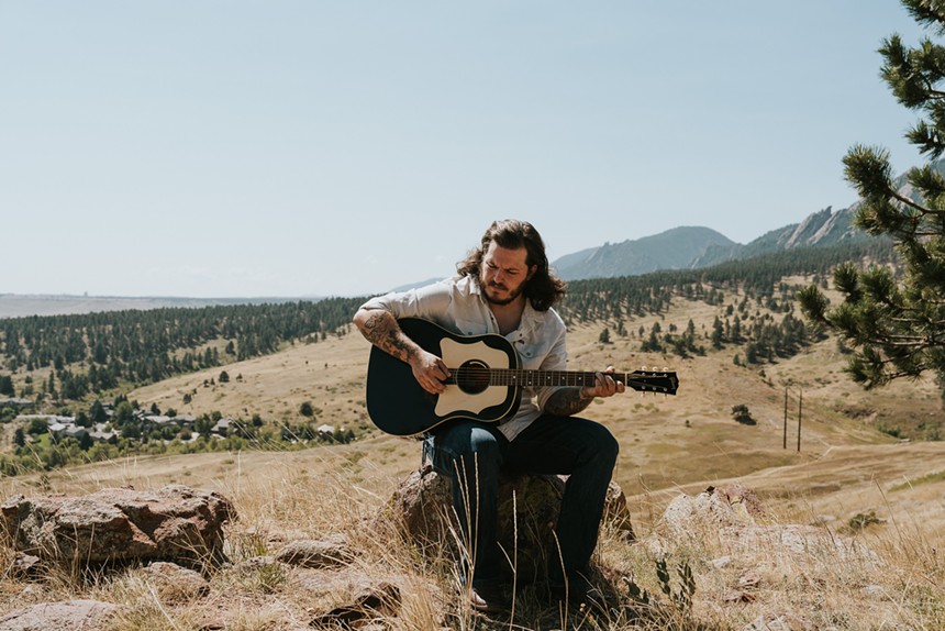 Danno Simpson sits on a rock playing guitar in the Boulder foothills with the Flatirons behind him.