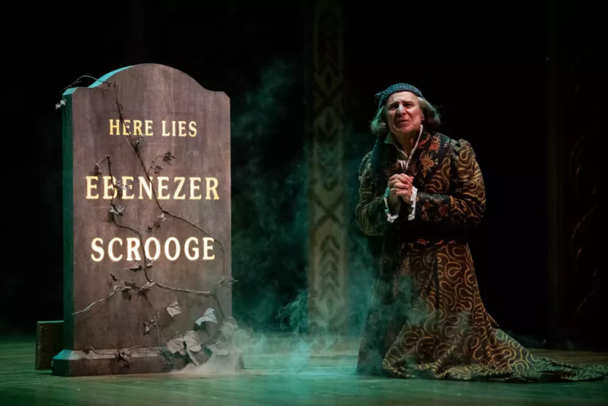 An old man kneels next to a tombstone that reads, "here lies Ebenezer Scrooge."