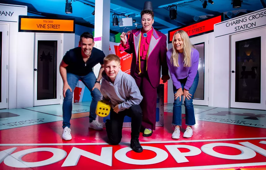 A boy rolls a dice on a large Monopoly board, surrounded by supportive spectators.
