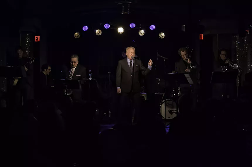 A man sings center stage, surrounded by a band.