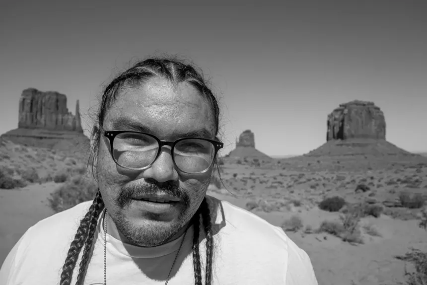 A black-and-white headshot of a man wearing glasses in the desert.