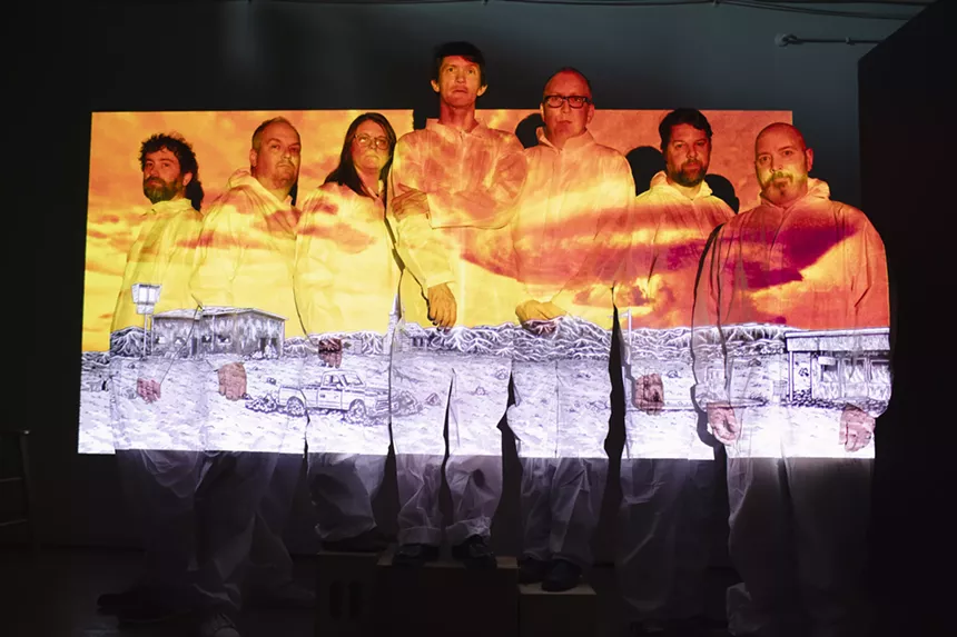 The members of Cursive stand lined against a wall with an image of a mushroom cloud against a fiery sky and a devastated landscape projected across their bodies.