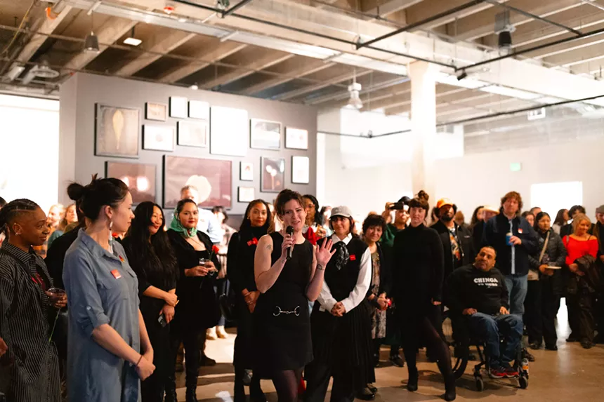 A large crowd listens to a woman speak into a microphone in an art gallery.