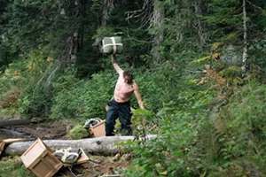 Ryan Willis standing on the forested shoreline, holding shelter-building supplies above his head