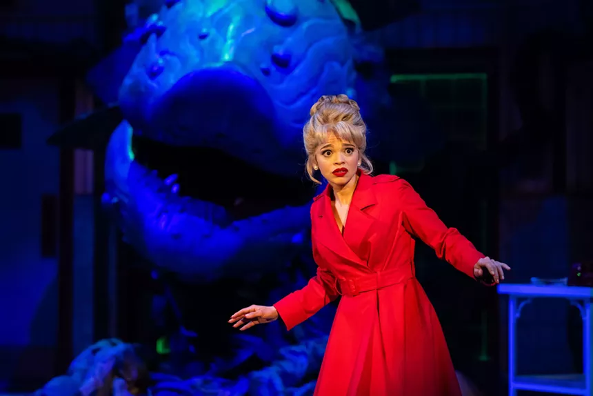 A woman in a red dress stands in front of a giant plant puppet.