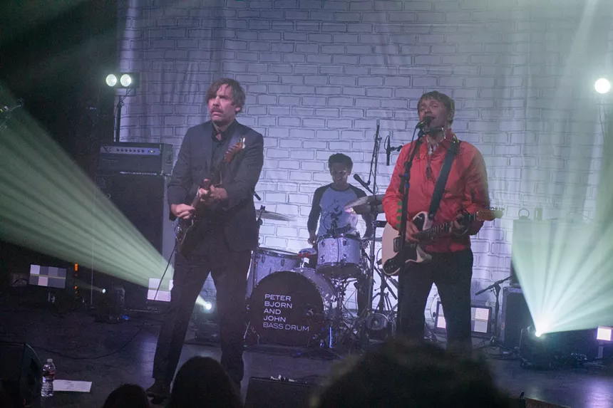Three band members singing and playing guitar, bass, and drums on stage