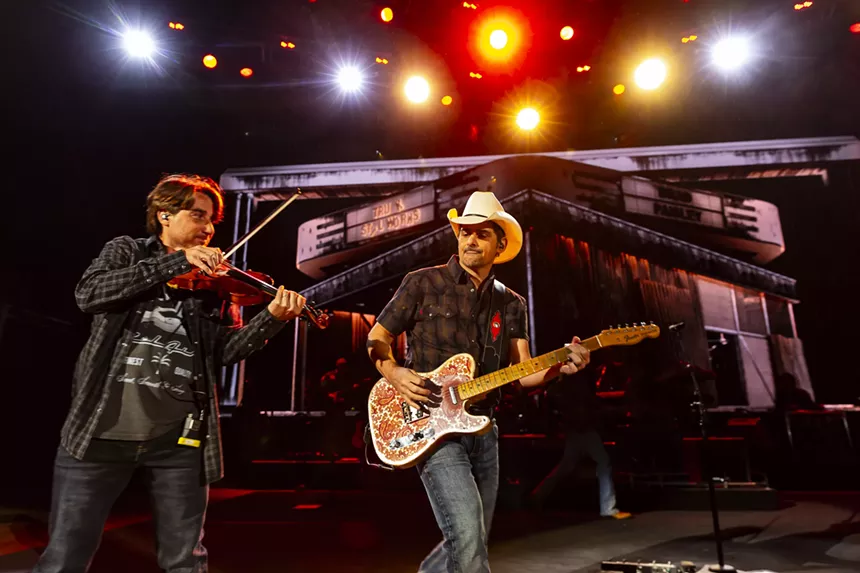 Brad Paisley on stage at Red Rocks with stage lights behind him and a violinist next to him.