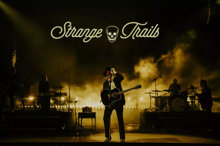 Lord Huron performing on stage at Red Rocks with fog behind him, and a stage sign labeled Strange Trails hangs above him.