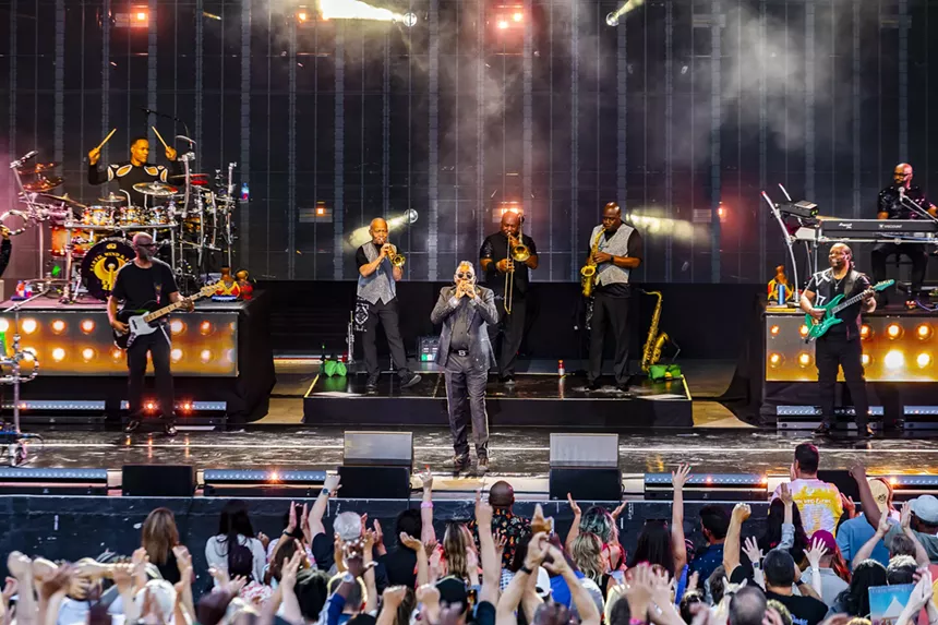 Members of Earth, Wind & Fire performing on stage with concert fans raising their hands in the air.
