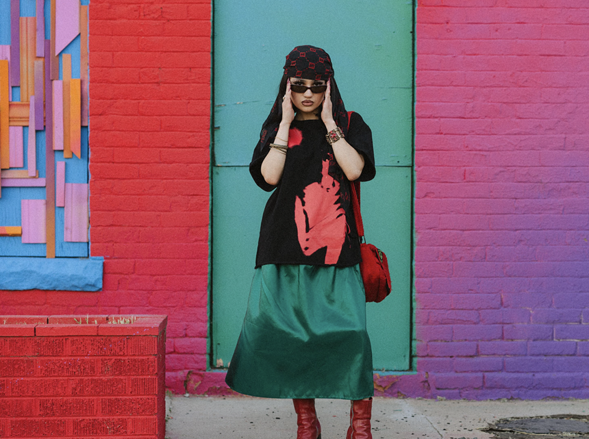 Kitty wears a mix of red, black and green clothing while standing in front od a red, green and pink painted wall.