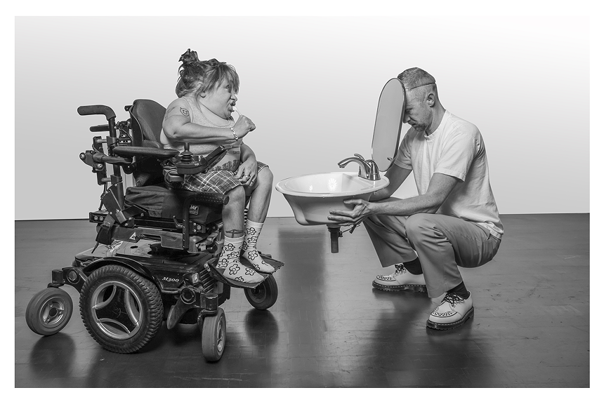 A woman in a wheelchair brushes her teeth while a squatting man holds a sink and mirror