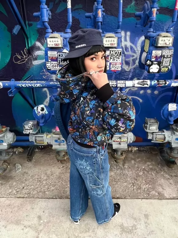Kitty wears a tom-boy-esque outfit while posing in front of top a spray painted wall with pipes.