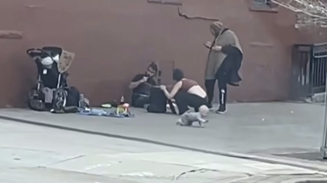 A baby crawling on a sidewalk while his mother and friends hang out nearby.