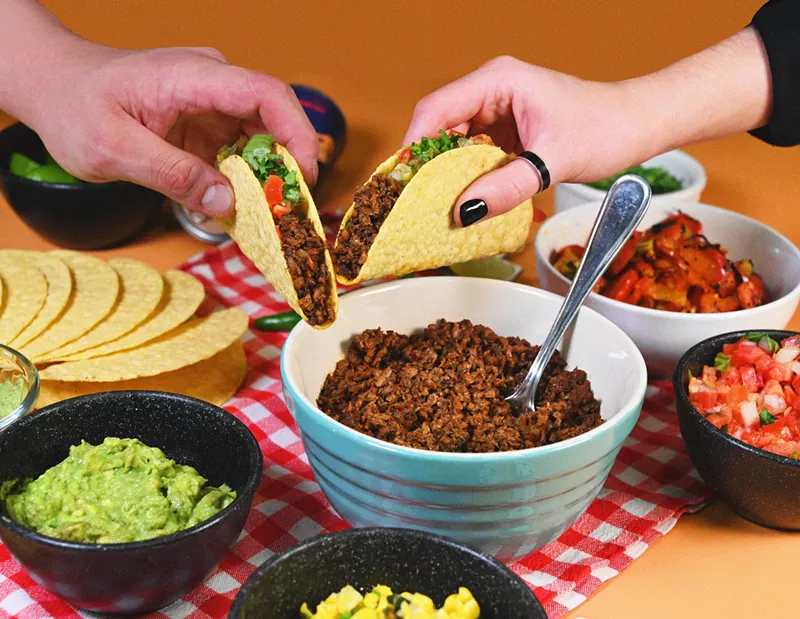 Image: Two hands holding hard shell tacos over bowls of Illegal Pete's menu items