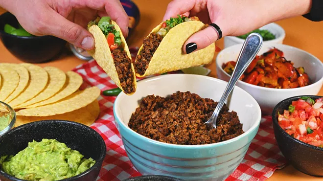 Two hands holding hard shell tacos over bowls of Illegal Pete's menu items